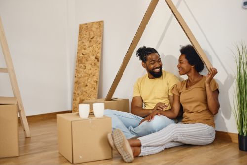A Black heterosexual couple sits smiling on the living room floor of their new home after moving in. There are boxes on the hardwood floor around them. The woman jokingly holds a piece of cardboard that arches over the couple imitating a roof over their heads.