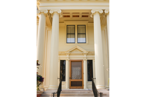 A closeup view of a Greek Revival architecture house. The large front porch is straddled by tall white columns that reach up to the second story of the home.