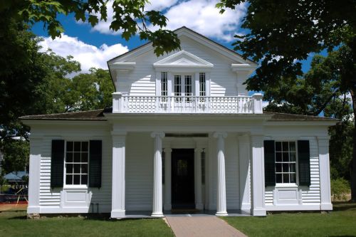 A street-level view of an example of Greek Revival architecture. It is a white house with columns in the front, a second-story balcony, windows with shutters, and an overall formal appearance.