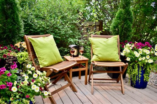 A private backyard patio where two wooden chairs with light green pillows are set up among flowers in ceramic planters, a lantern with a candle in it, and trees all around, which hide the fence in the background.