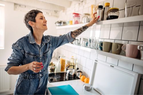 A White woman with short greying hair reaches for kitchen items on her open shelves above her countertops.