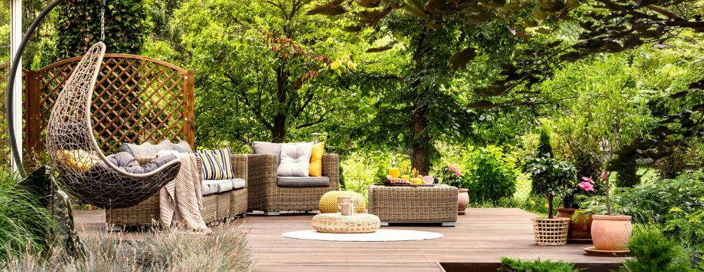 A private backyard patio with a hanging chair, comfortable wicker patio furniture, a refinished wooden deck, and lush greenery surrounding the seating area.