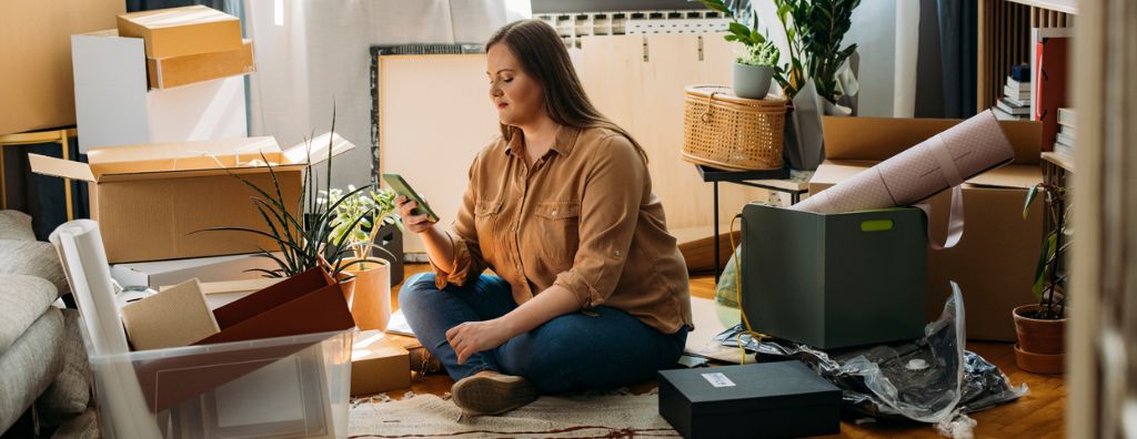 A young White woman looks at her checklist on her phone as she sits on the living room floor of her new home after moving in. Her belongings are scattered around the room. Some things are still in boxes, and others are unpacked.