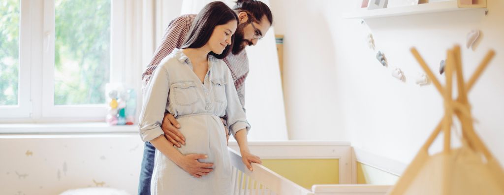 A young man and pregnant woman prepare for their DIY nursery décor project as they look over a crib in a playroom.