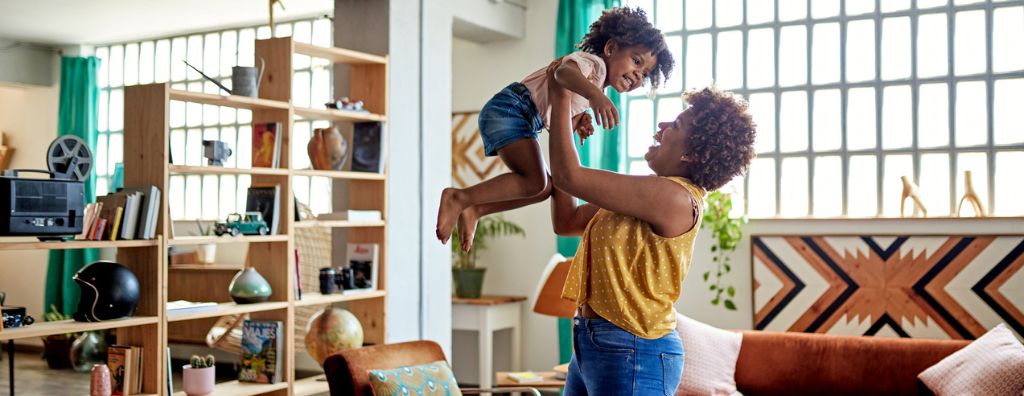 A Black mother lifts her young daughter in the air of her apartment. The decorations show a well-designed small space, with elements of multifunctional design throughout the open living room.