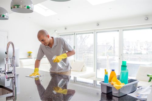 A man is cleaning his kitchen stovetop. He is wearing orange rubber gloves and scrubbing soap over the surface as he works to keep a tidy home.
