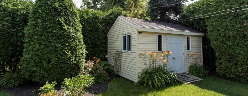 A yellow backyard shed ADU surrounded by green hedges and flower beds with long yellow flowers in them.