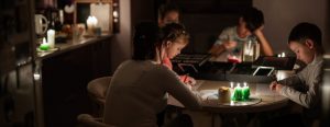 A mother and her four children sit at the dining room table during a power outage. They have lit candles to provide light and are drawing pictures together.