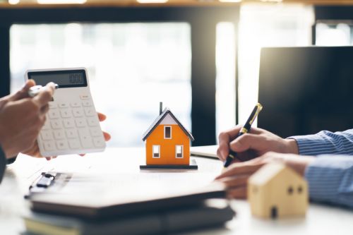 A close up shot of hands across a table from one another in an office. On the left, the hands of the real estate agent calculate fair market value on a calculator. On the right, the hands of the client are writing notes with a pen.