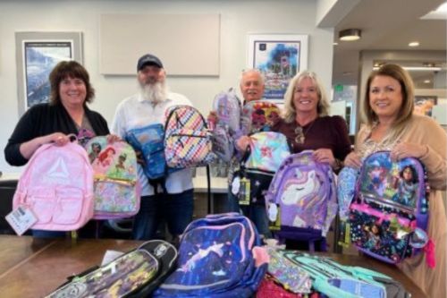 A group of staff from Windermere Camano Island hold up backpacks at a backpack drive for local schoolchildren.