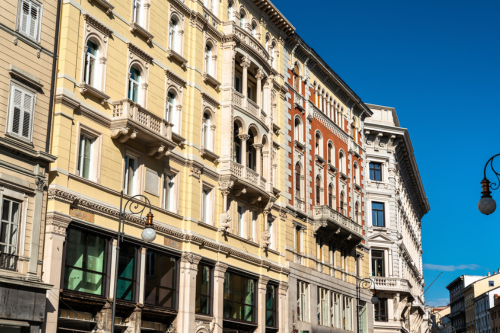 An example of Italianate architecture: an extended row house structure in downtown Trieste, Italy. Each room has a curved window decorated with ornate trim facing the street and a small ledge. There are columns on the ground floor and halfway up the central building.