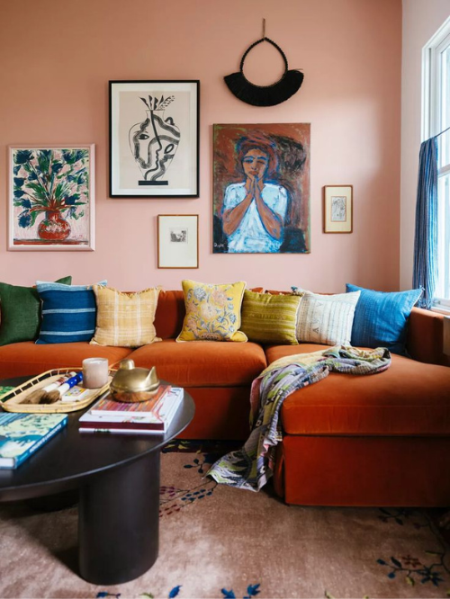 A living room with a colorful palette of jewel tones with a bright orange couch, yellow and blue throw pillow, and black and gold accents includes a Peach Fuzz colored accent wall with a gallery of art displayed.