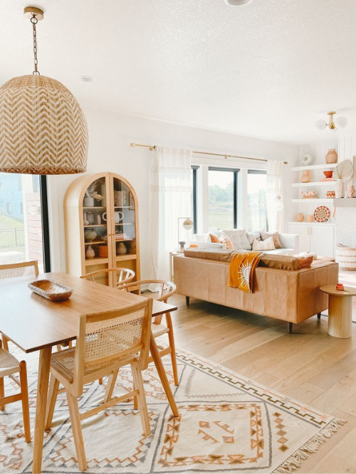 A living room with a neutral palette of white, natural wood, and browns includes orange hued accents in the rug, throw pillows, and displayed décor on the shelves. 