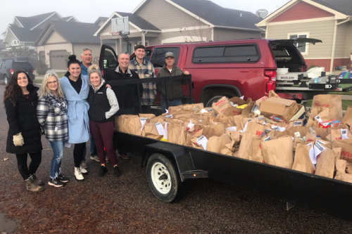 A group of Windermere agents pose smiling by paper bags full of food from their annual Can the Cats Food Drive benefiting Missoula Food Bank and Community Center. 