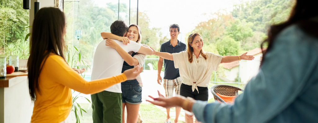 Small group of friends arriving at a party, meeting and greeting host. Two people are hugging and others have their arms open ready for a hug.