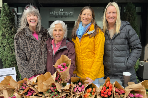 Four white women stand, smiling, behind a table with tulip bouquets wrapped in brown paper. They’re in rain jackets and they stand in front of the Windermere Spokane office, with the Windermere logo displayed on the wall behind them. 
