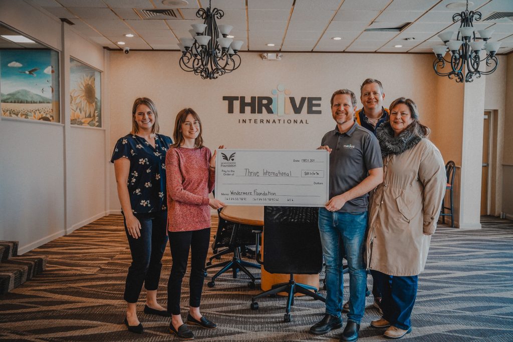 Five white people pose smiling with a giant check in a boardroom. The Thrive International logo is on the wall behind them. 