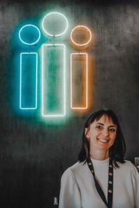 Yuliia smiles in front of a lit neon sign in the shape of the Thrive International logo. 