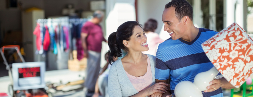 Happy heterosexual couple walks away from a yard sale blurred in the background with a lamp in his hands.