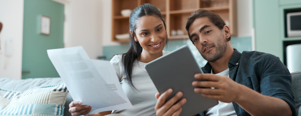 Heterosexual couple looks over documents printed in her hands and on his tablet. They’re smiling together on their couch in their rental home as they see that their savings have built up enough over time to afford a down payment for their first home.