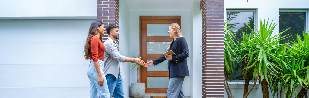 Real estate agent showing a couple a new house. The house is contemporary. All are happy and smiling and shaking hands. The couple are casually dressed and the agent is in a suit. The house has a brick facade